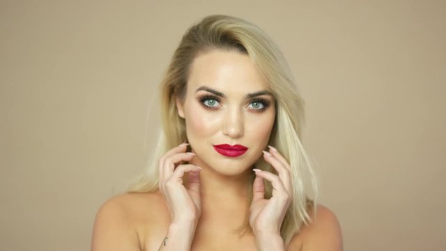 Portrait of pretty young woman with blonde hair and red lips touching head and looking at camera in studio.