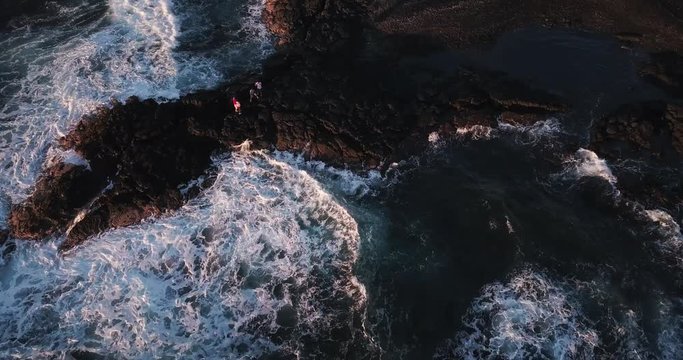 Hawaii drone aerial static looking down at waves hitting black sand volcanic beach as people take photos on the edge