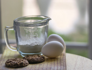 A glass measurement cup, two eggs and baked brown chocolate cookies on top of a wooden board, with soft light and out of focus background