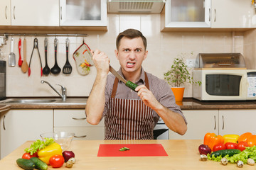 Angry crazy caucasian young man in apron, brown shirt sitting at table, attack cuts vegetable for salad with knife in light kitchen. Dieting concept. Healthy lifestyle. Cooking at home. Prepare food.