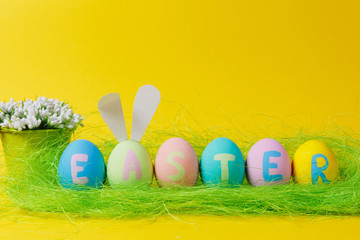 Row of six colorful pastel monophonic painted Easter eggs with inscription Easter, fun bunny ears, white flowers in bucket in green grass isolated on yellow background. Copy space for advertisement.