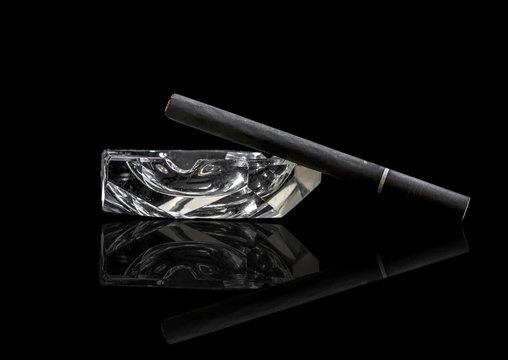 Single Kretek black clove cigarrete on a glass ashtray, black background and reflective surface.
