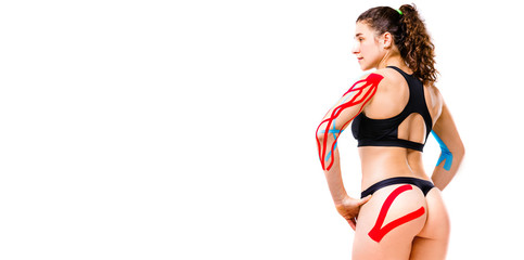 sexy female athlete with beautiful booty stands half sideways on white background in black top and bikini.On body is pasted kinesiology tape.Colored tapes for treatment of muscles,tendons in athletes