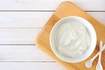 Greek yogurt in a white bowl, above view on a natural wood cutting board against white wood.