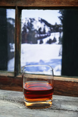 glass of whiskey or other alcohol in front of a cabin window on a cold winter day with trees and mountains in the view