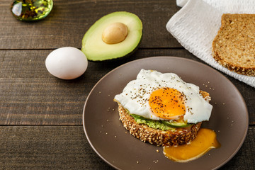 sandwich with avocado and a fried egg on a brown plate on a rustic wood and table bread on white kitchen towel.