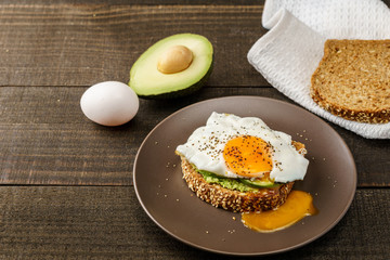 sandwich with avocado and a fried egg on a brown plate on a rustic wood and table bread on white kitchen towel.