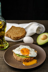 sandwich with avocado and a fried egg on a brown plate on a rustic wood and table bread on white kitchen towel.