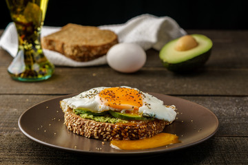 sandwich with avocado and a fried egg on a brown plate on a rustic wood and table bread on white kitchen towel.