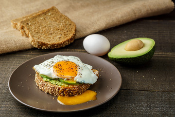 sandwich with avocado and a fried egg on a brown plate on a rustic wood and table bread on sackcloth.