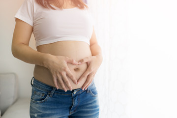 Pregnant Asian Woman holding her hands in a heart shape on her belly near window at home.copy space.