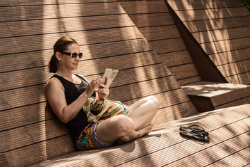 Woman sitting at wood bench in shade, using smartphone, touching touchscreen;