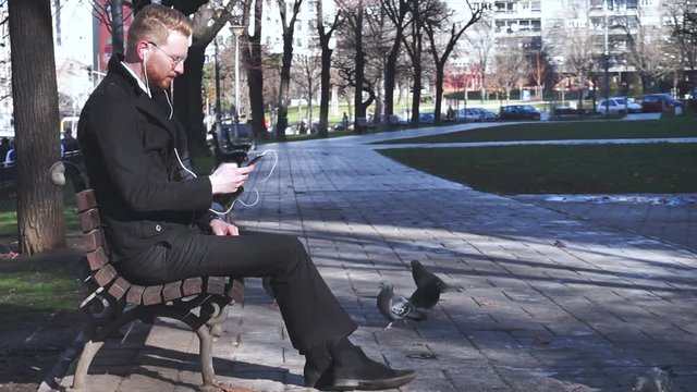 Young ginger businessman sitting alone in park and checking his social media on phone while listening to music via white headphones
