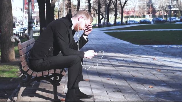 Sad young ginger businessman siting alone in park and checking his social media while listening to sad music via white headphones