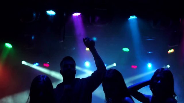 The silhouette of a cheerful friends dancing in the nightclub in the dark with the light colored spotlights, slow motion.