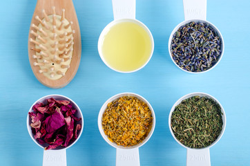 Wooden hairbrush and plastic scoops with olive oil, dry marigold, lavender, parsley and dog rose flowers. Aromatherapy, herbal medicine and natural skin and hair care concept. Top view, copy space. 