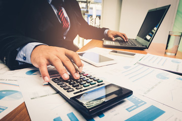 A businessman analyzing investment charts at his workplace and using his laptop and touch calculator.