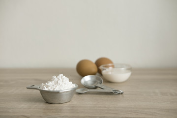 BAKERY INGREDIENTS
Bakery ingredients, white flour in a measuring cup, milk and eggs in transparent cups with measuring spoon are on a wood table.