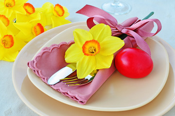Simple table setting arrangement with cream porcelain dishes, red Easter egg, yellow daffodil flower, cutlery and napkin