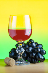 A glass of red wine next to bunch of black grapes and a cork on a wooden table on a yellow green gradient background (concept)