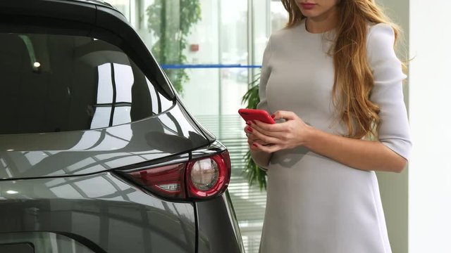 Cropped shot of a long haired woman standing near a new auto at the dealership using her smart phone typing messaging communication mobility consumerism driving buying.