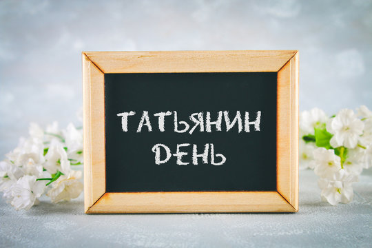The inscription in Russian: Tatyanin day. Russian holiday on student's day. A chalkboard is surrounded by white flowers on a gray background.