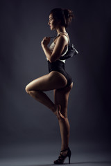 Fototapeta na wymiar Portrait of young smiling female dancer in black high waist panties and white top standing on one leg in high-heeled shoe, another leg is barefoot and raised. She is holding a boot behind her shoulder