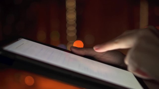 Close-up of woman hands using tablet computer touchscreen outdoors in city at nignt. Searching for information online on internet browser pages. Tiny snowflakes are falling on the screen.