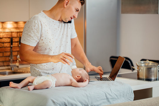 Dad Trying To Work, Cooking And Talk On Phone While Standing With His Newborn Babe In Home Office Interior