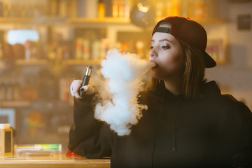 Young pretty woman in red cap smoke an electronic cigarette at the vape shop. Hip-hop style....