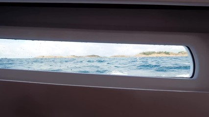 Speed boat is moving rapidly across the sea, creating waves. Large foam waves beat against the ship side. In the background a horizon and a mountain line. Rectangular porthole in a speed boat