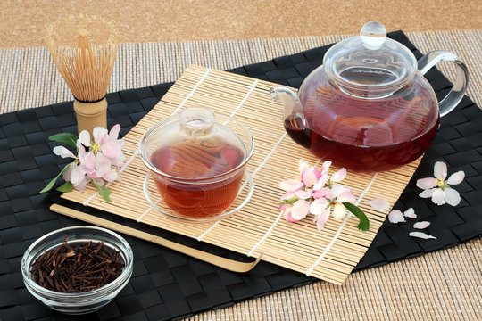 Kuchika Japanese roasted green twig tea with glass teapot and cups, dried twigs, stirrer, whisk and blossom flowers on bamboo background. Has many health benefits.