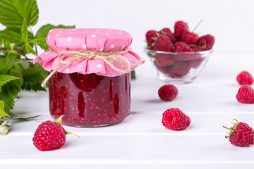 Raspberry jam in glass jar, fresh  ripe raspberry and green leaves on white wooden table