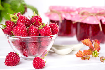 Red fresh raspberries in a glass bowl and Raspberry jam with green leaves on white background.