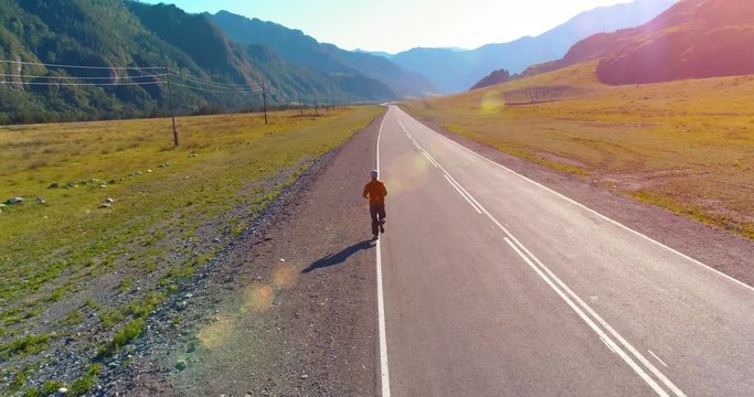 UHD 4K aerial view. Low altitude flight behind of sporty man at perfect asphalt road. Sunrise in mountain. Green meadow and sun rays on horizon. Fast horizontal movement.