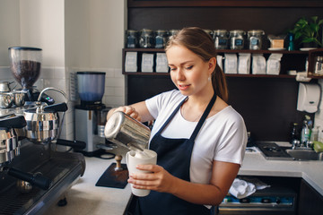 Portrait of smiling young Caucasian woman barista  in apron pouring milk in coffee. Business owner concept.
