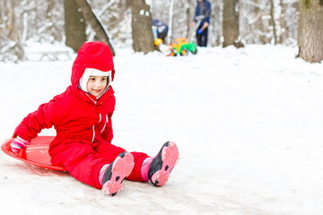 Fototapeta na wymiar Pretty smiling little girl in her ski suit sliding down a small snow covered hill with her sledge