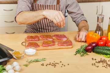 Close up young man in apron sitting at table with vegetables, cooking at home preparing meat stake from pork, beef or lamb, salt meat, in light kitchen with wooden surface, full of fancy kitchenware.
