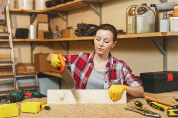 Beautiful smiling caucasian young brown-hair woman in plaid shirt, gray T-shirt, yellow gloves sawing piece of wood with saw, working in carpentry workshop at wooden table place with different tools.