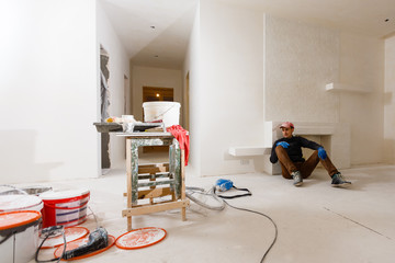 Handyman is having a rest leaned on a radiator and sitting on the floor with his tools young man
