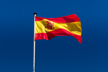 The flag of Spain, a horizontal tricolour of red, yellow and red, charged with the Spanish coat of arms against the blue sky, Spain