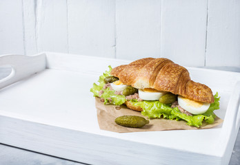 Croissant sandwich with tuna, hard boiled egg, salad and cucumber on white tray. Breakfast concept