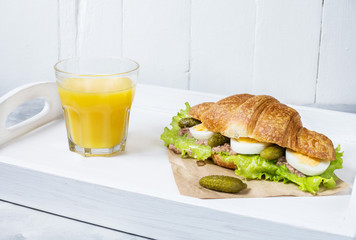 Croissant sandwich with tuna, hard boiled egg, salad and cucumber on white tray. Glass of orange juice. Breakfast