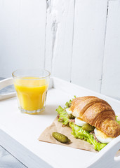 Croissant sandwich with tuna, hard boiled egg, salad and cucumber on white tray. Glass of orange juice. Breakfast concept