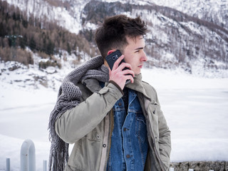 Fototapeta na wymiar Handsome young man making a call with smarpthone while standing at snowy mountain
