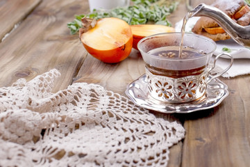 Homemade pastries with persimmons, for breakfast. Beautiful glass with tea. Kettle. Wooden table, tablecloth with lace. Free space for text or advertising.