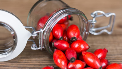 Spilled red rosehips on wood table. Close-up of lying glass jar and medicinal fruits of wild rose on the brown background.