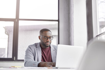 Fototapeta premium Indoor shot of professional black male freelancer works remotely on laptop computer, plans working schedule, keyboards information, focused into screen. Trader analyzes financial market online