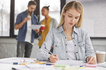 Smart blonde pretty female student wears denim jacket, writes down something in papers, prepares project work, drinks coffee from paper cup and her groupmates talk with each other in background