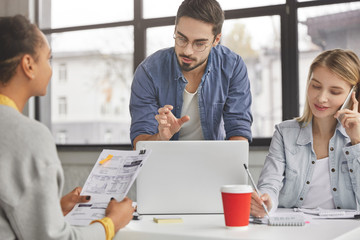 Fototapeta premium Attractive unshaven male coach speaks to his female colleagues. Concentrated young skilled marketing expert tries to present his advertising concept for trading company, gestures as explains chart.